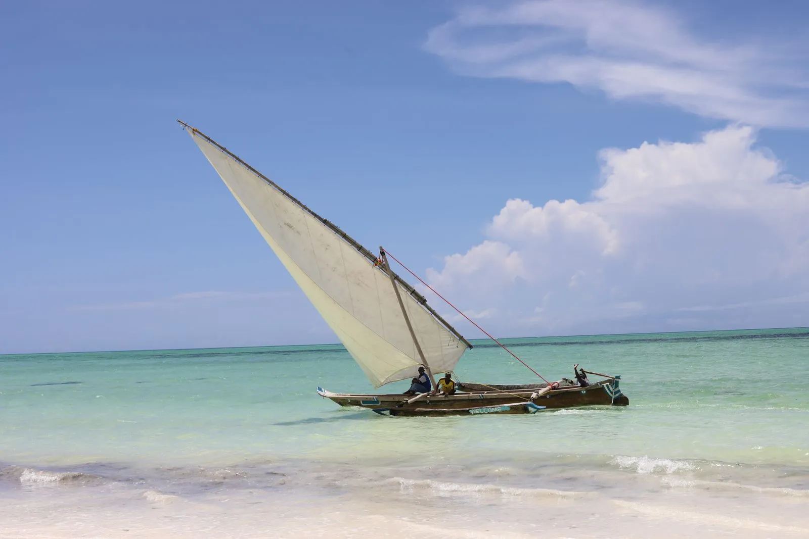 Dhow under a white sail in turquoise water off the beach
