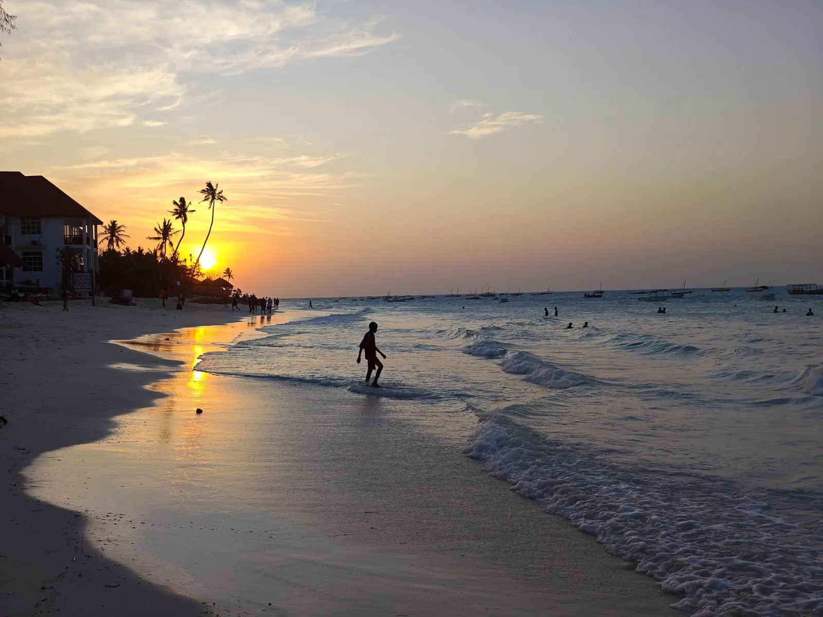 Sunset over the sea with a hotel and palms on the left and a lone silhouette in the surf