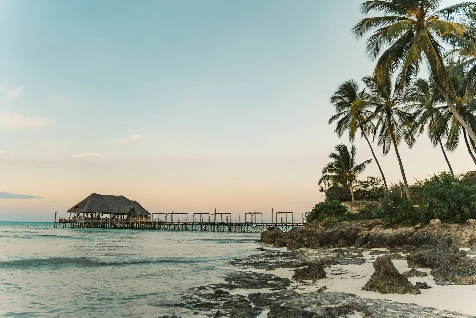 Long wooden jetty to a thatched pavilion over the sea, with palms and coral rock in front