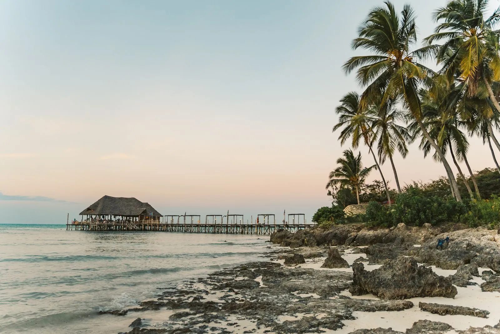 Wooden jetty to a thatched pavilion beside coral rocks and palms at dusk